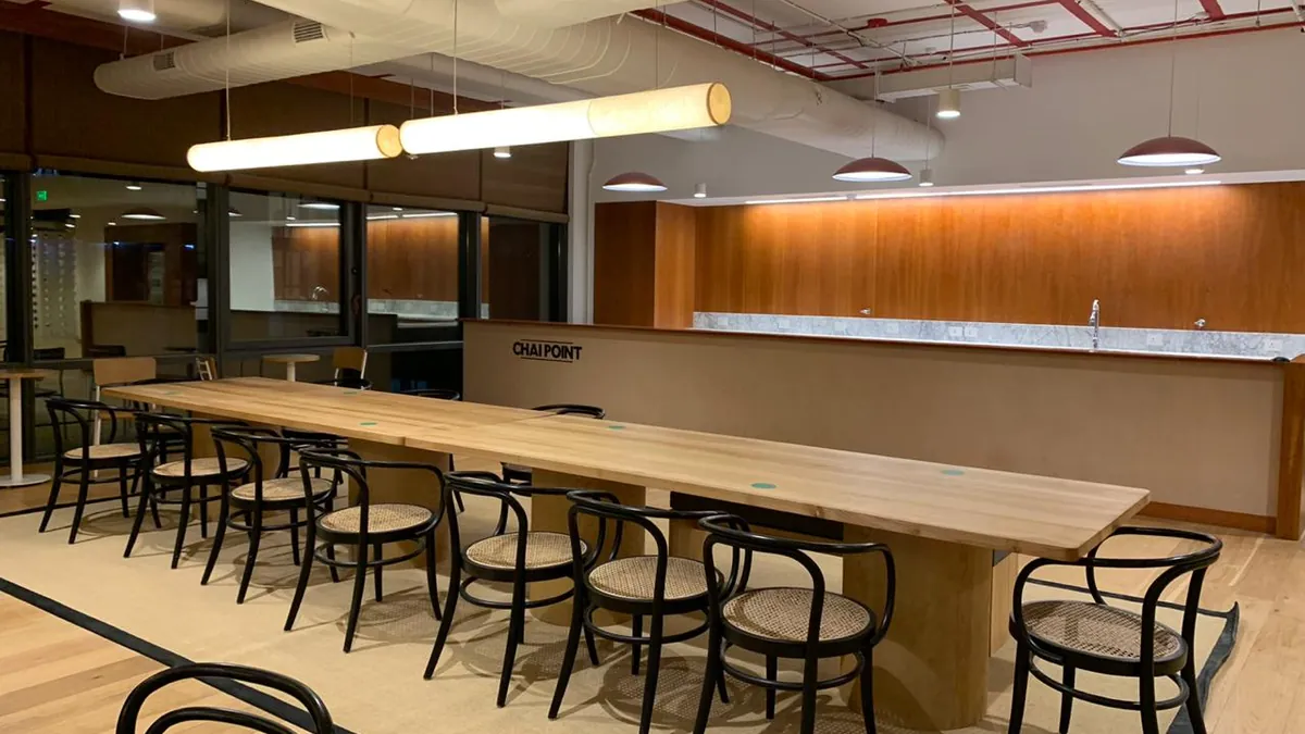 Office breakout area with a long solid timber table, cane-back chairs, timber wall panelling and exposed ceiling services